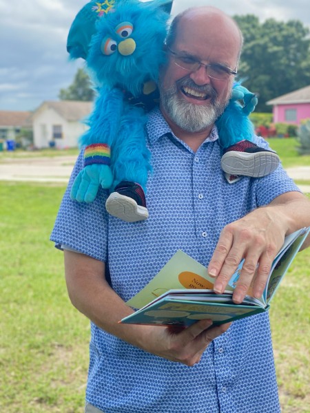 Thomas Kenny Smiling Reading a Book with a Blue Puppet on Shoulders
